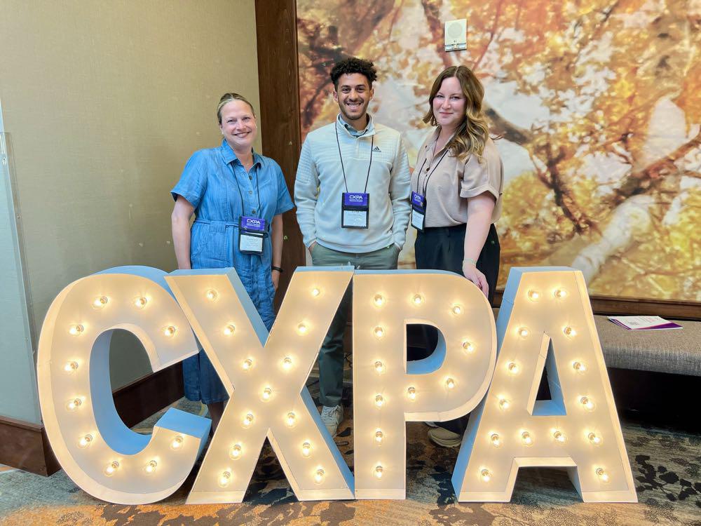Two women and a man standing behind marquee letters spelling CXPA